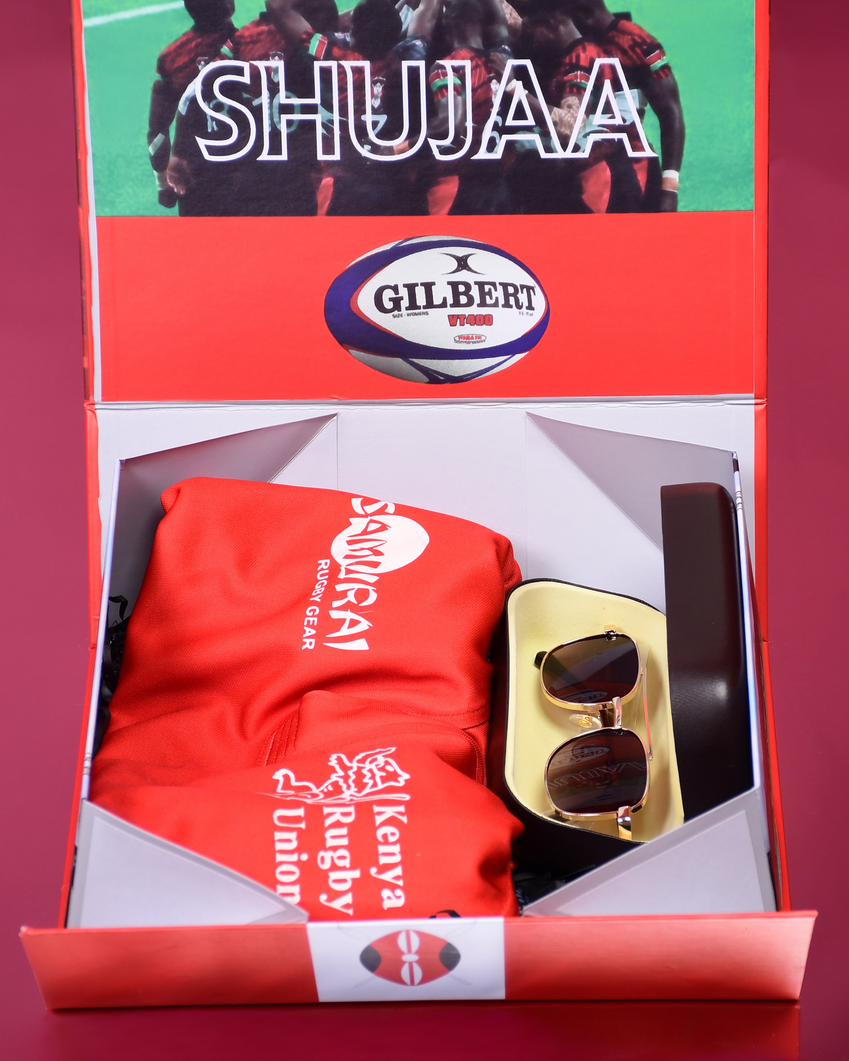 Box containing a red shirt, sunglasses, and a bottle labeled 'Shujaa' with 'Gilbert' branding.